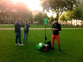 A person stands on a green lawn with a sign that says 'FREE SILENT DISCO' and 'HEADSETS'. Two other individuals interact nearby, wearing headphones. Trees and a building are in the background, with sunlight filtering through.