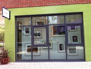 The image depicts a storefront with a predominantly lime green brick facade and large windows. Inside, there are various framed photographs or artworks visible through the windows. The store has a modern and artistic appearance.