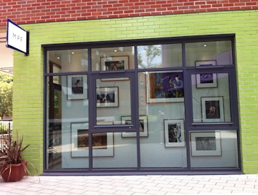 The image depicts a storefront with a predominantly lime green brick facade and large windows. Inside, there are various framed photographs or artworks visible through the windows. The store has a modern and artistic appearance.