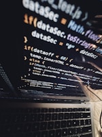 A close-up view of a laptop screen displaying lines of JavaScript code, with the keyboard partially visible at the bottom. The code is reflected on the screen, creating a layered visual effect. The environment appears dark, contributing to a focused atmosphere.