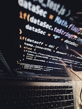 A close-up view of a laptop screen displaying lines of JavaScript code, with the keyboard partially visible at the bottom. The code is reflected on the screen, creating a layered visual effect. The environment appears dark, contributing to a focused atmosphere.