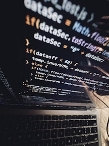 A close-up view of a laptop screen displaying lines of JavaScript code, with the keyboard partially visible at the bottom. The code is reflected on the screen, creating a layered visual effect. The environment appears dark, contributing to a focused atmosphere.