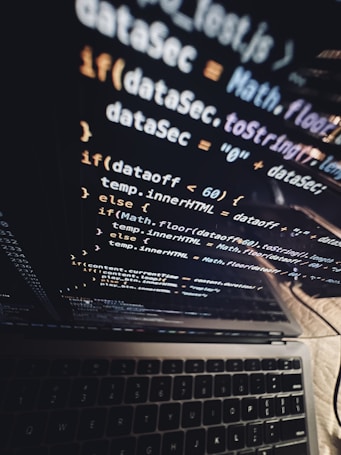A close-up view of a laptop screen displaying lines of JavaScript code, with the keyboard partially visible at the bottom. The code is reflected on the screen, creating a layered visual effect. The environment appears dark, contributing to a focused atmosphere.