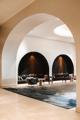 A stylish, modern lounge area with curved archways and black velvet seating. The space is accentuated by soft lighting that creates a warm ambiance. Circular tables and plush chairs add to the sophisticated design, while the tile flooring complements the contemporary decor.