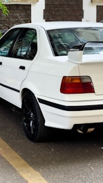 A white sports sedan parked on the street, featuring tinted windows and a prominent spoiler with a BMW Motorsport logo. The car has black alloy wheels and distinctive red and amber taillights. A stone wall with a green plant is visible in the background.