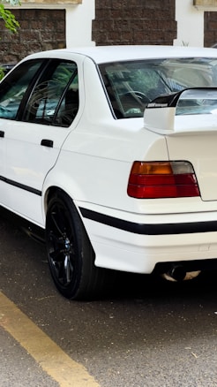 A white sports sedan parked on the street, featuring tinted windows and a prominent spoiler with a BMW Motorsport logo. The car has black alloy wheels and distinctive red and amber taillights. A stone wall with a green plant is visible in the background.