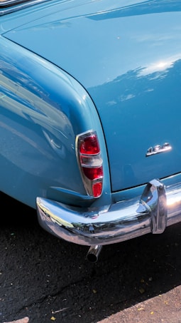 A vintage car features a prominent blue paint job with shiny chrome accents. The tail light is reflective and partially red, situated next to a large chrome bumper. The car model emblem is visible on the rear. The pavement below is dark and slightly textured, with a few small leaves scattered.