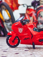 A small red toy motorcycle with a firefighting symbol on the front part is placed on a surface with a terrazzo pattern. In the background, there are blurred images of bicycles with bright colors.