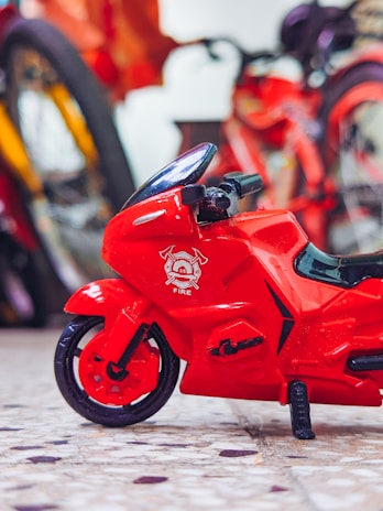 A small red toy motorcycle with a firefighting symbol on the front part is placed on a surface with a terrazzo pattern. In the background, there are blurred images of bicycles with bright colors.