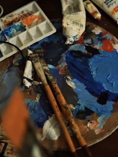 A painter's workspace featuring a palette covered with various shades of blue and some red paint. There are several brushes, some with paint on their bristles, and tubes of paint in the background. An open paint palette shows remnants of mixed colors.