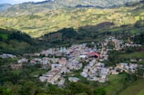 A small village nestled in a lush, mountainous landscape with rolling hills and dense forests. The village features a mix of colorful buildings, including a prominent structure with a red roof, and is crisscrossed by narrow, winding roads.