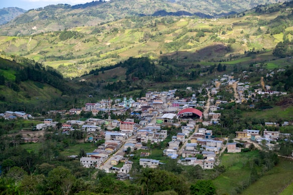 A small village nestled in a lush, mountainous landscape with rolling hills and dense forests. The village features a mix of colorful buildings, including a prominent structure with a red roof, and is crisscrossed by narrow, winding roads.