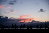 A serene evening scene with silhouettes of people seated in chairs by the water. The sky is beautifully painted with hues of blue, gray, and a striking pink from the setting sun, creating a calm and peaceful ambiance. Clouds scatter across the sky, adding texture and depth to the image.