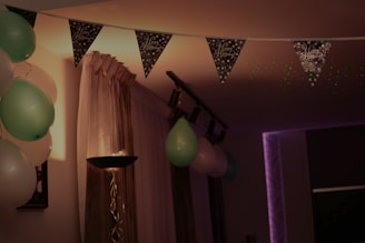 A room decorated for a celebration with a string of 'Happy Birthday' pennant banners and colorful balloons, mostly in green and white. There is a standing lamp with a unique, twisted design and soft lighting. Curtains cover a window, creating a cozy and intimate ambiance.