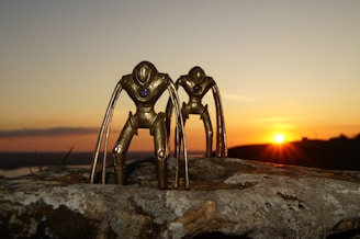 Two metallic, abstract sculptures resembling humanoid figures with elongated limbs stand on a large rock surface. The sculptures have a futuristic design and feature a small purple gem or sphere embedded in their chests. A vivid sunset with an orange and pink sky forms the background, casting warm light over the scene.