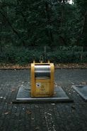 A yellow recycling bin with signage stands on a cobblestone path, surrounded by dense green foliage and trees in the background. The bin appears to be in a park or wooded area.