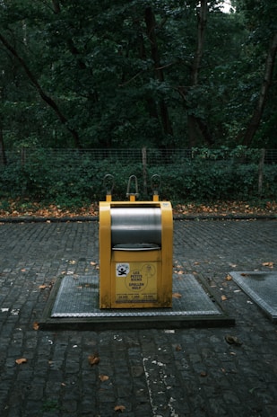 A yellow recycling bin with signage stands on a cobblestone path, surrounded by dense green foliage and trees in the background. The bin appears to be in a park or wooded area.
