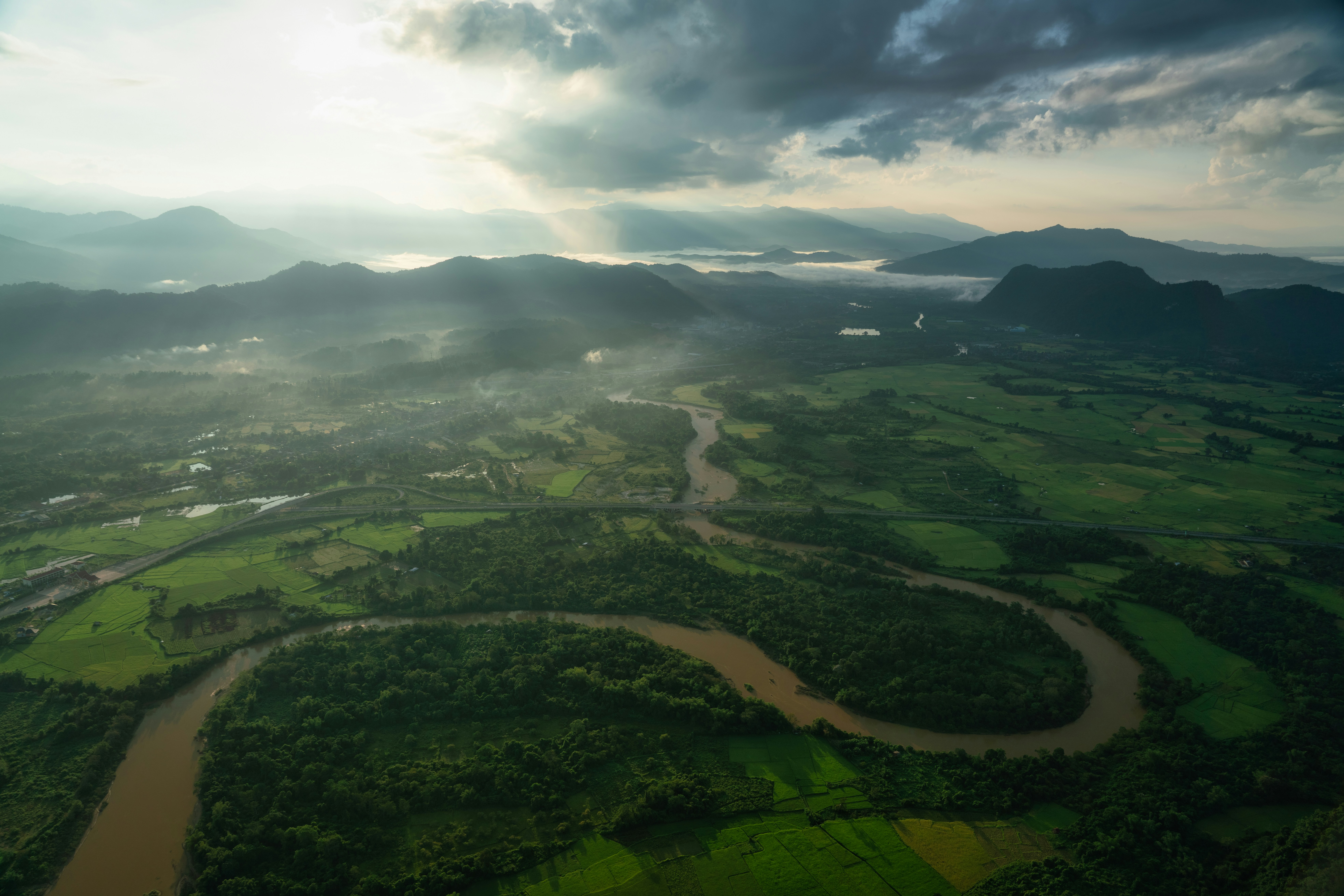 an aerial view of a river running through a lush green valley, Captivating landscape of Vang Vieng, Laos, showcasing the majestic mountains enveloped in a serene mist. This image embodies the raw beauty and tranquility of Southeast Asia