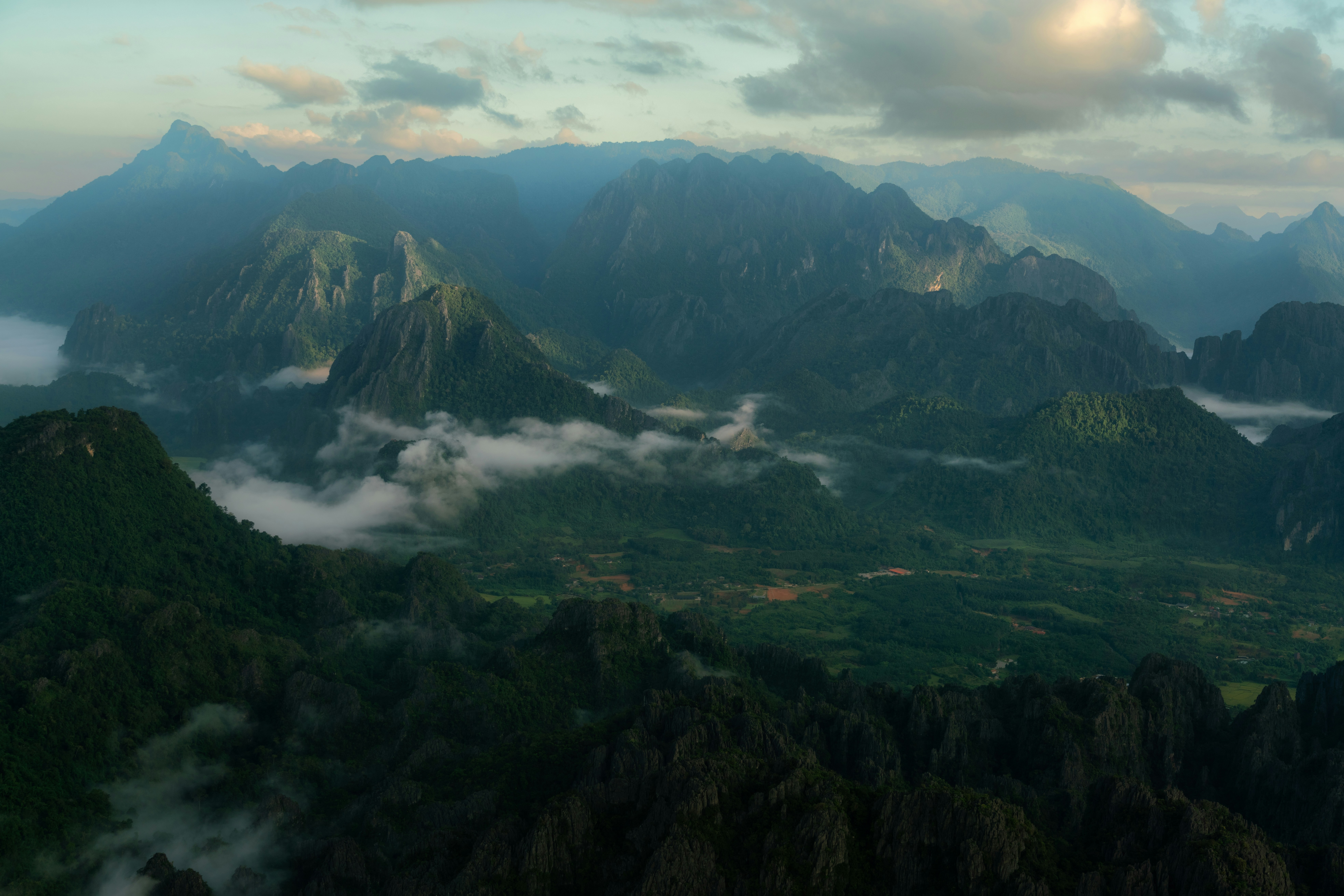 an aerial view of a mountain range with low lying clouds, Captivating landscape of Vang Vieng, Laos, showcasing the majestic mountains enveloped in a serene mist. This image embodies the raw beauty and tranquility of Southeast Asia