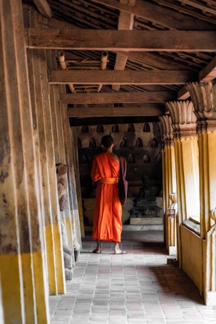 A monk in a vibrant orange robe stands in a narrow corridor of an ancient temple, surrounded by wooden beams and old stone walls. The corridor is dimly lit, and the atmosphere is peaceful and contemplative, with small Buddha statues visible in the background.