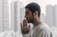 A profile of a person with their hands together as if in prayer, with multiple tattoos visible on the neck and hands. In the background, tall, blurred buildings are visible, suggesting an urban setting. The person wears a light-colored long-sleeve shirt.