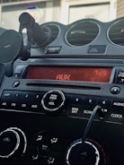 A car dashboard featuring a digital display reading 'AUX' and '7:25'. Below the screen are control buttons for various functions including DISP, AM, FM, CD, and AUX. A power and volume knob is prominently located in the center. Air vents and a phone holder are visible, and a cord is plugged into an auxiliary input.