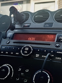 A car dashboard featuring a digital display reading 'AUX' and '7:25'. Below the screen are control buttons for various functions including DISP, AM, FM, CD, and AUX. A power and volume knob is prominently located in the center. Air vents and a phone holder are visible, and a cord is plugged into an auxiliary input.