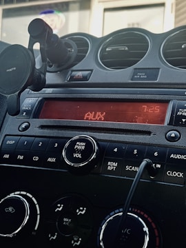 A car dashboard featuring a digital display reading 'AUX' and '7:25'. Below the screen are control buttons for various functions including DISP, AM, FM, CD, and AUX. A power and volume knob is prominently located in the center. Air vents and a phone holder are visible, and a cord is plugged into an auxiliary input.