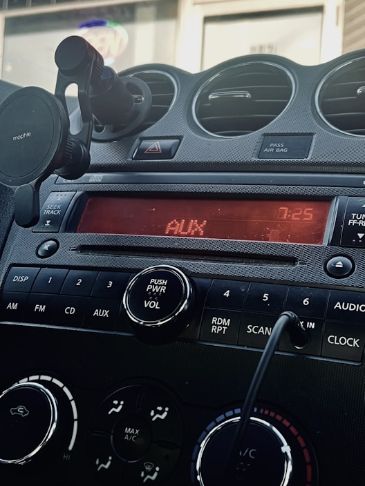 A car dashboard featuring a digital display reading 'AUX' and '7:25'. Below the screen are control buttons for various functions including DISP, AM, FM, CD, and AUX. A power and volume knob is prominently located in the center. Air vents and a phone holder are visible, and a cord is plugged into an auxiliary input.
