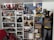 A room crammed with various pop culture collectibles and figures, primarily focused around science fiction and superhero genres. There are several shelves filled with action figures, models, books, and DVDs. Notable items include a Darth Vader helmet, Baby Yoda figure, and numerous Doctor Who memorabilia. Posters from movies like Back to the Future and Batman adorn the walls. A mannequin dressed as Batman, with a facehugger from Alien attached to its chest, stands next to the bookshelves.