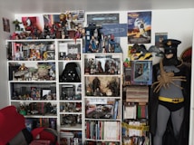 A room crammed with various pop culture collectibles and figures, primarily focused around science fiction and superhero genres. There are several shelves filled with action figures, models, books, and DVDs. Notable items include a Darth Vader helmet, Baby Yoda figure, and numerous Doctor Who memorabilia. Posters from movies like Back to the Future and Batman adorn the walls. A mannequin dressed as Batman, with a facehugger from Alien attached to its chest, stands next to the bookshelves.
