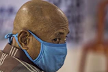 A close-up of a bald person wearing a blue face mask, looking sideways. The background is blurred, drawing focus to the person's head and mask.