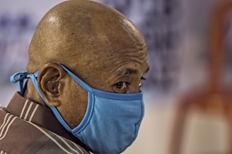 A close-up of a bald person wearing a blue face mask, looking sideways. The background is blurred, drawing focus to the person's head and mask.