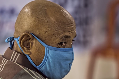 A close-up of a bald person wearing a blue face mask, looking sideways. The background is blurred, drawing focus to the person's head and mask.