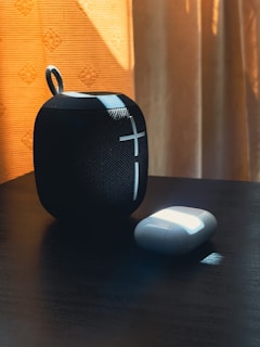 A small, portable speaker with a mesh texture stands next to wireless earbuds in a charging case. Sunlight casts soft shadows and highlights on the objects, creating a contrast with the dark wooden surface and warm, textured curtains in the background.