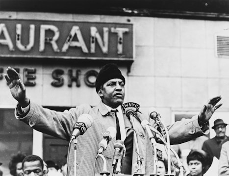 A man wearing a coat and hat is standing behind a cluster of microphones, delivering a speech in front of a crowd. There are several people gathered, some with serious expressions. The background features a building with visible signage.