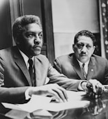 Two men in suits sit at a table, appearing engaged and focused on an ongoing discussion or meeting. Papers and documents are present on the table, indicating a formal setting.