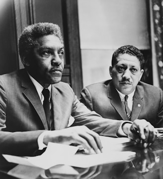 Two men in suits sit at a table, appearing engaged and focused on an ongoing discussion or meeting. Papers and documents are present on the table, indicating a formal setting.