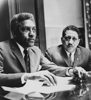 Two men in suits sit at a table, appearing engaged and focused on an ongoing discussion or meeting. Papers and documents are present on the table, indicating a formal setting.