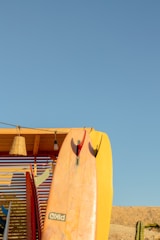 A collection of surfboards is propped against a wooden structure beneath a clear blue sky. One prominent surfboard in the center has a mix of pink and yellow hues. A wicker lamp and a light bulb hang from the wooden overhang. In the background, there's a sandy terrain with a cactus on the right side.
