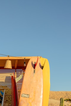 A collection of surfboards is propped against a wooden structure beneath a clear blue sky. One prominent surfboard in the center has a mix of pink and yellow hues. A wicker lamp and a light bulb hang from the wooden overhang. In the background, there's a sandy terrain with a cactus on the right side.