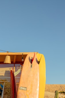 A collection of surfboards is propped against a wooden structure beneath a clear blue sky. One prominent surfboard in the center has a mix of pink and yellow hues. A wicker lamp and a light bulb hang from the wooden overhang. In the background, there's a sandy terrain with a cactus on the right side.