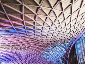 The image displays an architectural ceiling structure with a diamond-shaped grid pattern. The design features a combination of metal beams and glass panels, illuminated with gradient lighting in shades of pink, purple, and blue. The modern, geometric pattern stretches across the space, creating a dynamic and visually striking effect.