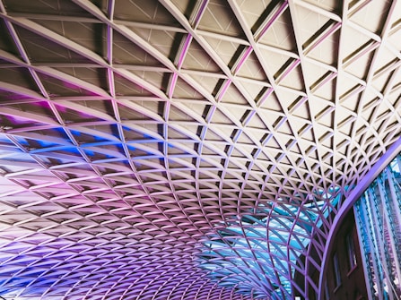 The image displays an architectural ceiling structure with a diamond-shaped grid pattern. The design features a combination of metal beams and glass panels, illuminated with gradient lighting in shades of pink, purple, and blue. The modern, geometric pattern stretches across the space, creating a dynamic and visually striking effect.