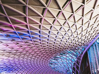 The image displays an architectural ceiling structure with a diamond-shaped grid pattern. The design features a combination of metal beams and glass panels, illuminated with gradient lighting in shades of pink, purple, and blue. The modern, geometric pattern stretches across the space, creating a dynamic and visually striking effect.