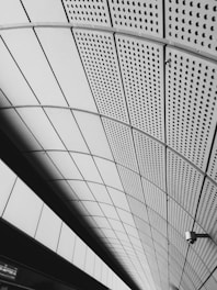 A geometric ceiling structure with a pattern of perforated panels creating a sense of depth and symmetry. The arrangement of shapes forms a modern architectural design, enhanced by the monochromatic color scheme. A surveillance camera is mounted on the right side, adding an element of security.