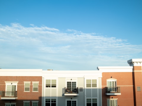 A modern apartment building with a clean, symmetrical facade features red brick and white paneling. Numerous rectangular windows line the structure, some with small balconies. The sky is bright and clear with a few wispy clouds.