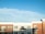 A modern apartment building with a clean, symmetrical facade features red brick and white paneling. Numerous rectangular windows line the structure, some with small balconies. The sky is bright and clear with a few wispy clouds.
