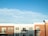 A modern apartment building with a clean, symmetrical facade features red brick and white paneling. Numerous rectangular windows line the structure, some with small balconies. The sky is bright and clear with a few wispy clouds.