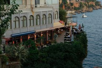 An outdoor restaurant terrace is nestled beside a building, overlooking a scenic coastline. Lush green plants surround the area, enhancing the serene ambiance. Several tables with umbrellas are arranged on the terrace, where people are dining and enjoying the seaside view. In the background, there is a calm body of water with a small boat, against a backdrop of cliffs and rustic buildings.